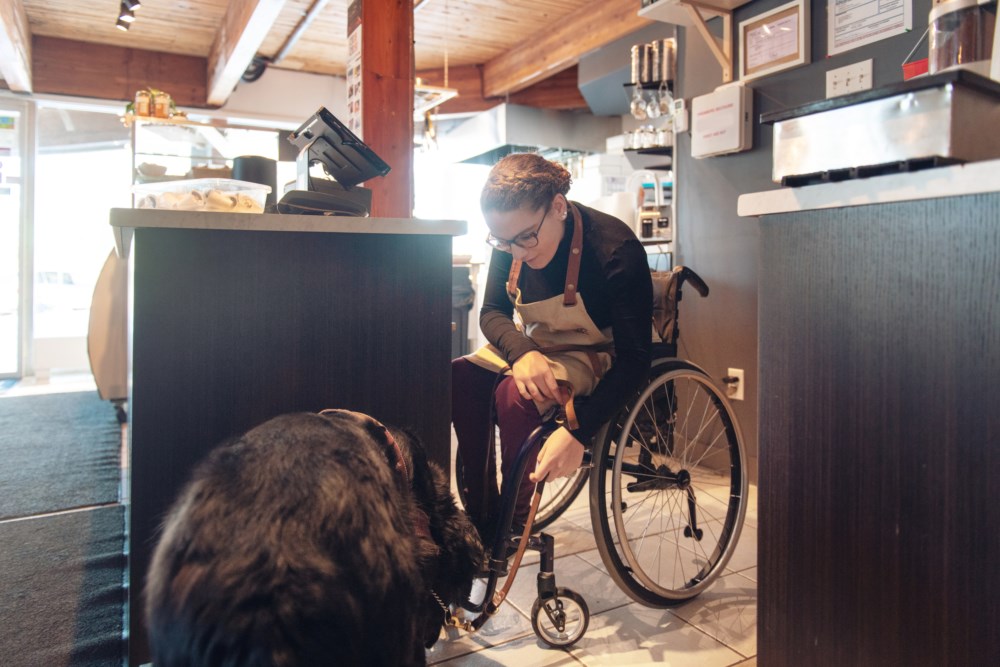 En kvinne i rullestol jobber som kasserer i en kafé. Hun smiler mens hun stryker en svart hund som står foran disken, og omgivelsene inkluderer en kaffemaskin og ulike matvarer bak disken.