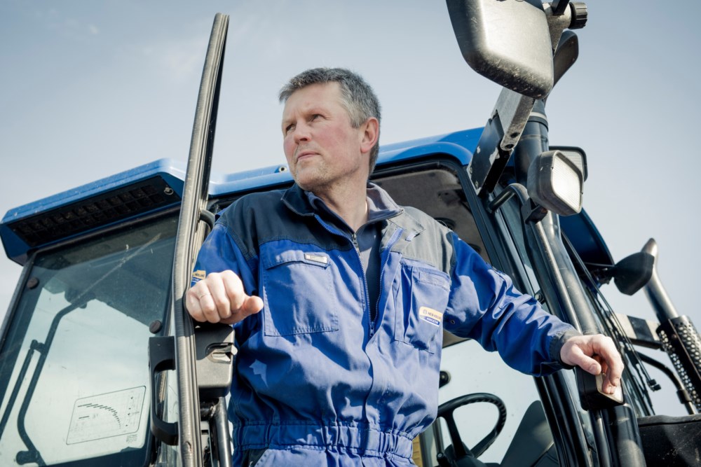 En mann i blå arbeidsdrakt med alvorlig uttrykk stiger ut av en traktor, med hånden på åpen dør og blikket rettet utover gården.